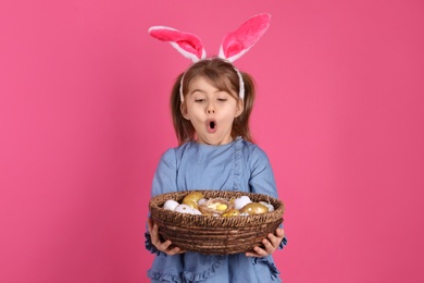 Photo of Surprised little girl with bunny ears holding wicker basket full of Easter eggs on pink background