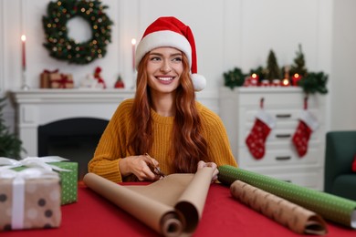 Photo of Beautiful young woman in Santa hat cutting wrapping paper at table. Decorating Christmas present