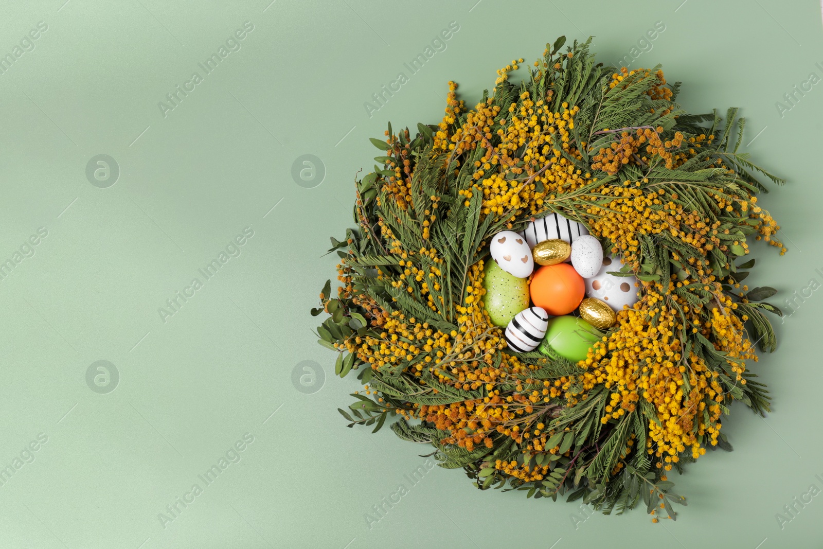 Photo of Festively decorated Easter eggs in nest of beautiful spring flowers on pale green background, top view. Space for text