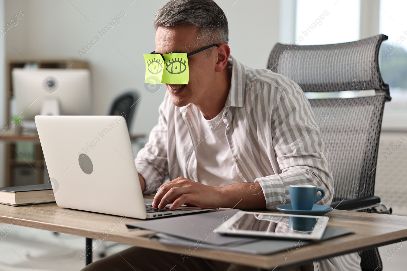 Photo of Man with fake eyes painted on sticky notes using laptop in office