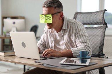 Man with fake eyes painted on sticky notes using laptop in office