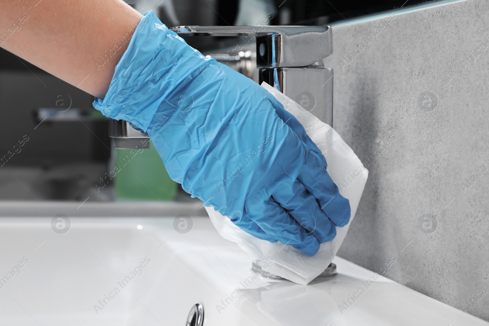 Photo of Woman in glove cleaning faucet of bathroom sink with paper towel, closeup