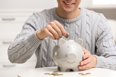 Photo of Man putting coin into piggy bank at white table, closeup