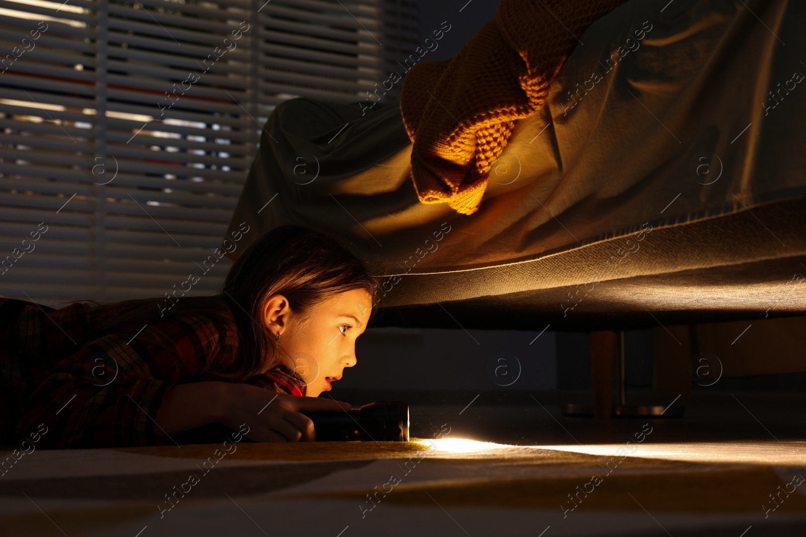 Photo of Little girl with flashlight looking for monster under bed at night