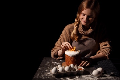 Photo of Young woman decorating traditional Easter cake at table against black background. Space for text