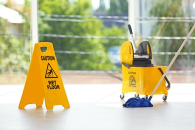 Photo of Safety sign with phrase Caution wet floor and mop bucket, indoors. Cleaning service