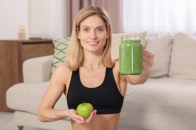 Photo of Young woman in sportswear with mason jar of fresh smoothie and apple at home