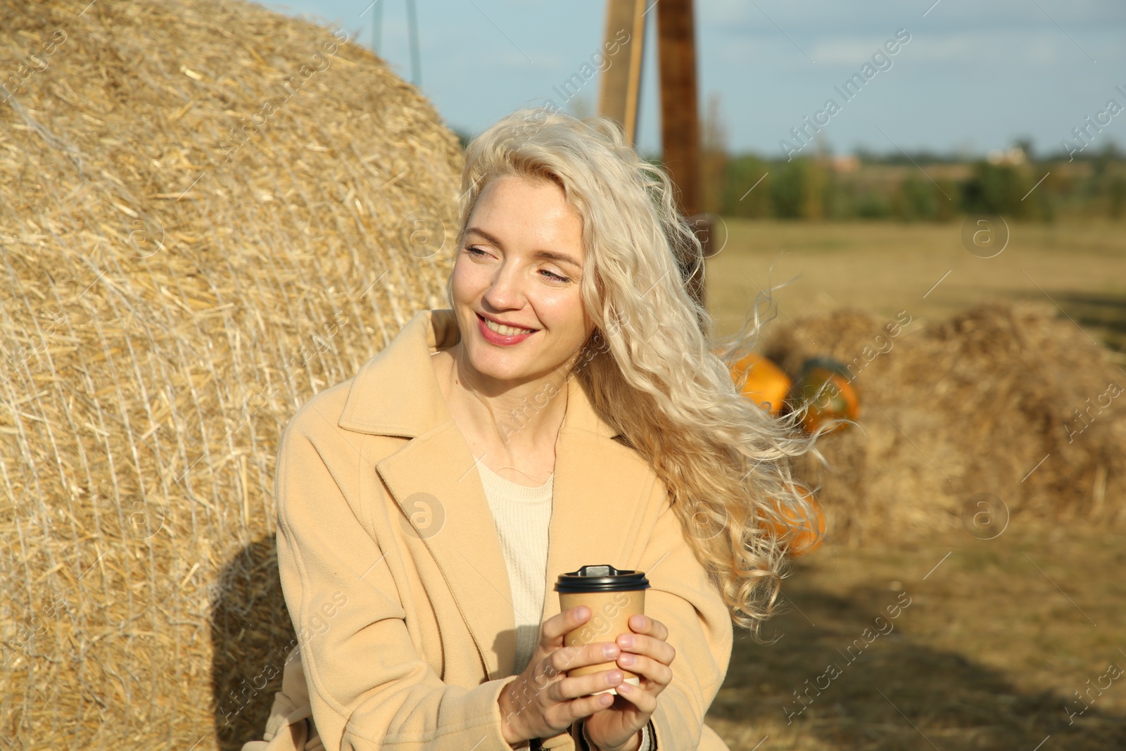 Photo of Beautiful woman with cup of hot drink sitting near hay bale outdoors, space for text. Autumn season