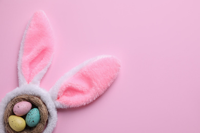 Headband with Easter bunny ears and dyed eggs in nest on pink background, flat lay. Space for text