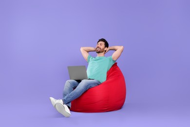 Photo of Happy man with laptop sitting on beanbag chair against purple background