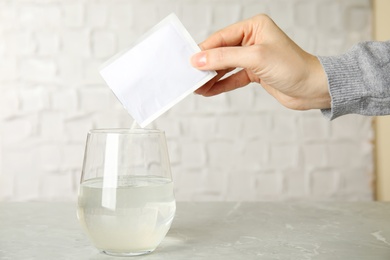Photo of Woman pouring powder from medicine sachet into glass of water on grey marble table, closeup