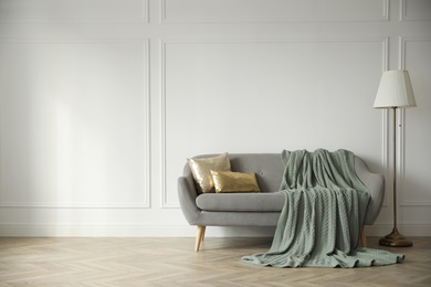 Photo of Stylish room interior with comfortable sofa, knitted blanket and lamp near white wall. Space for text