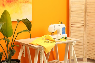 Photo of Modern sewing machine with cloth and craft accessories on wooden table near orange wall in room