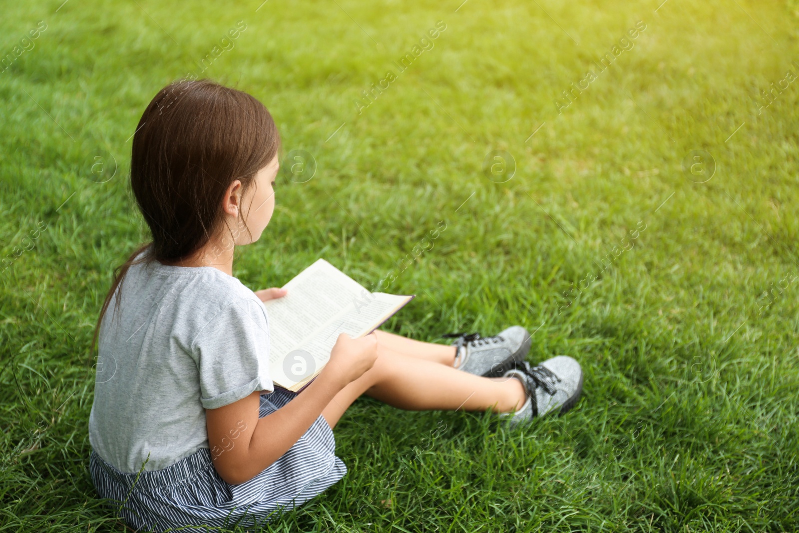 Photo of Cute little girl reading book on green grass in park