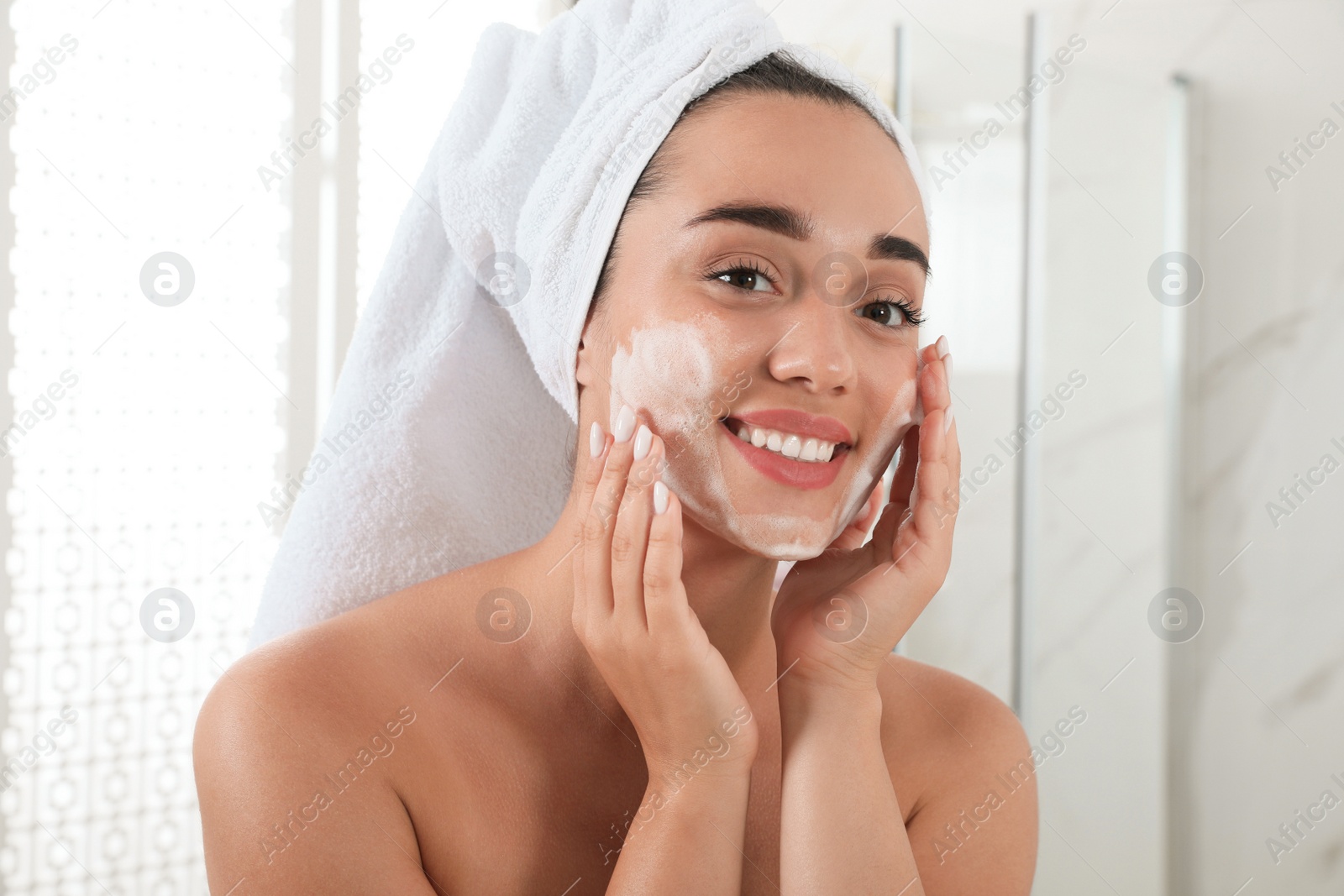 Photo of Beautiful young woman applying cleansing foam onto face in bathroom. Skin care cosmetic