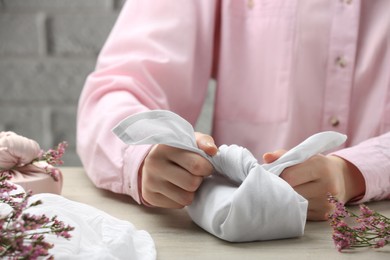 Furoshiki technique. Woman wrapping gift in white fabric at wooden table, closeup