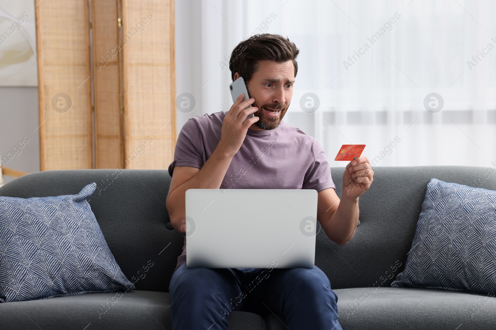 Photo of Emotional man with credit card and laptop talking on phone at home. Be careful - fraud