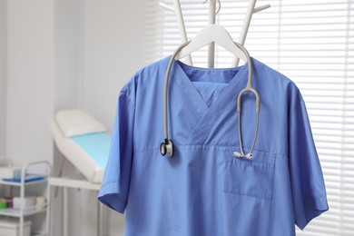 Photo of Blue medical uniform and stethoscope hanging on rack in clinic, closeup