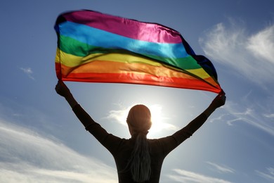 Photo of Woman holding bright LGBT flag against blue sky on sunny day, back view