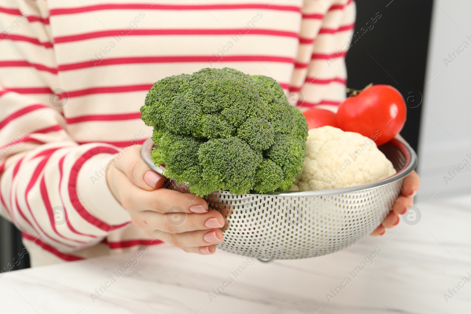 Photo of Woman holding colander with fresh vegetables at white marble table, closeup