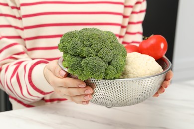 Woman holding colander with fresh vegetables at white marble table, closeup