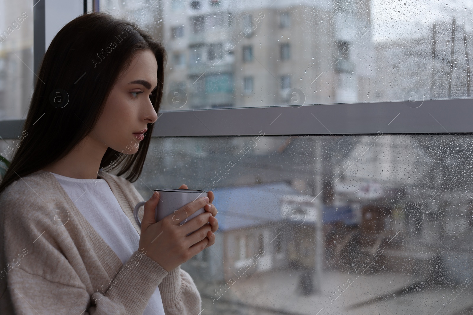 Photo of Melancholic young woman with drink looking out of window on rainy day, space for text. Loneliness concept