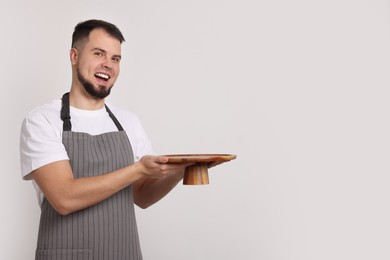 Photo of Happy professional confectioner in apron holding empty cake stand on light grey background. Space for text