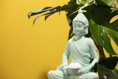 Buddhism religion. Decorative Buddha statue with burning candle and monstera against yellow wall, space for text