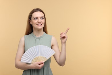 Happy woman with hand fan pointing on beige background, space for text