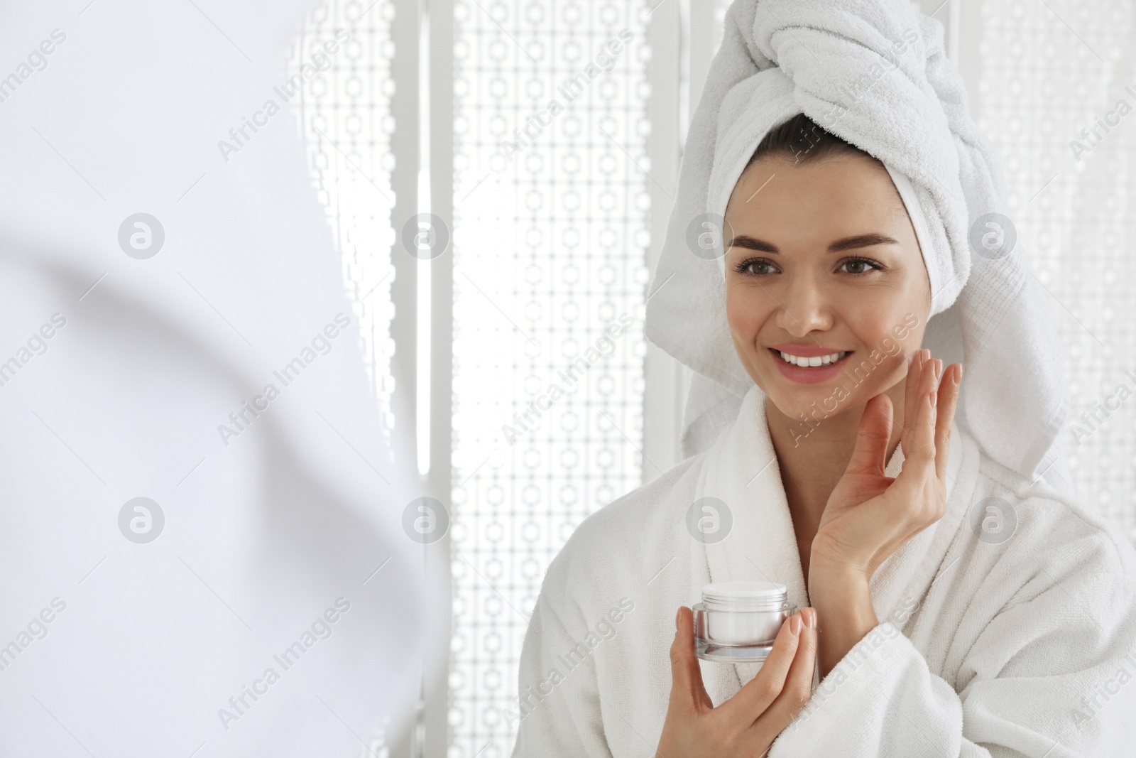 Photo of Beautiful young woman with hair wrapped in towel applying cream near mirror indoors