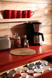 Photo of Open drawer with cups and coffeemaker near wooden wall indoors