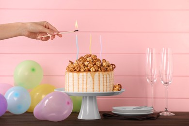 Woman lighting candle on caramel drip cake decorated with popcorn and pretzels at wooden table, closeup