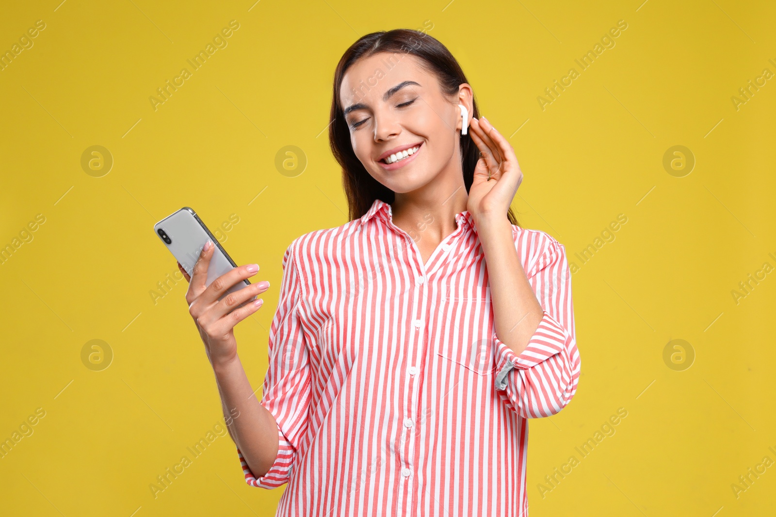 Photo of Happy young woman with smartphone listening to music through wireless earphones on yellow background