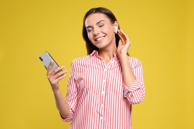 Happy young woman with smartphone listening to music through wireless earphones on yellow background