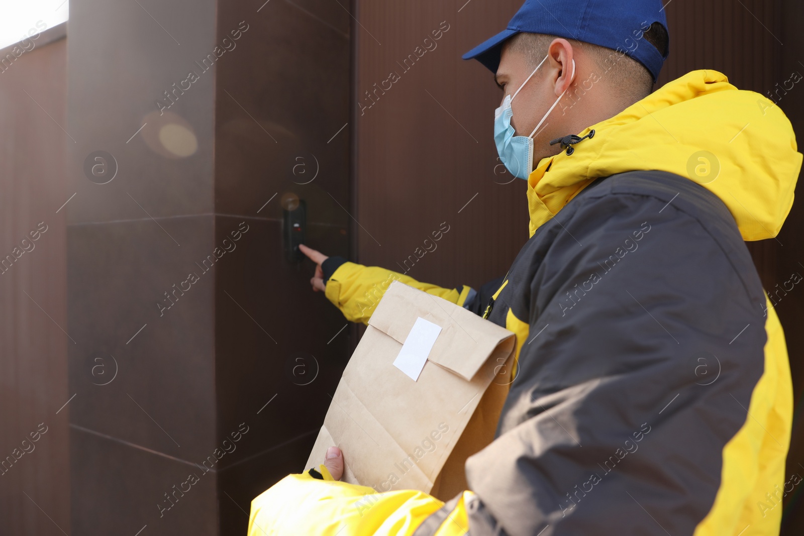 Photo of Courier in medical mask holding paper bag with takeaway food and ringing doorbell outdoors. Delivery service during quarantine due to Covid-19 outbreak
