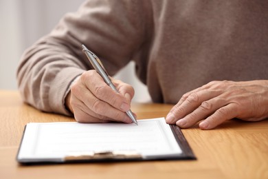 Photo of Senior man signing Last Will and Testament at wooden table indoors, closeup