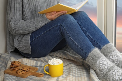 Photo of Woman with cup of hot cocoa reading book on window sill, closeup. Winter drink