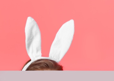 Little girl wearing headband with Easter bunny ears hiding behind table against color background, space for text