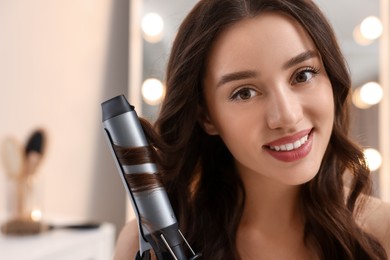 Smiling woman using curling hair iron indoors