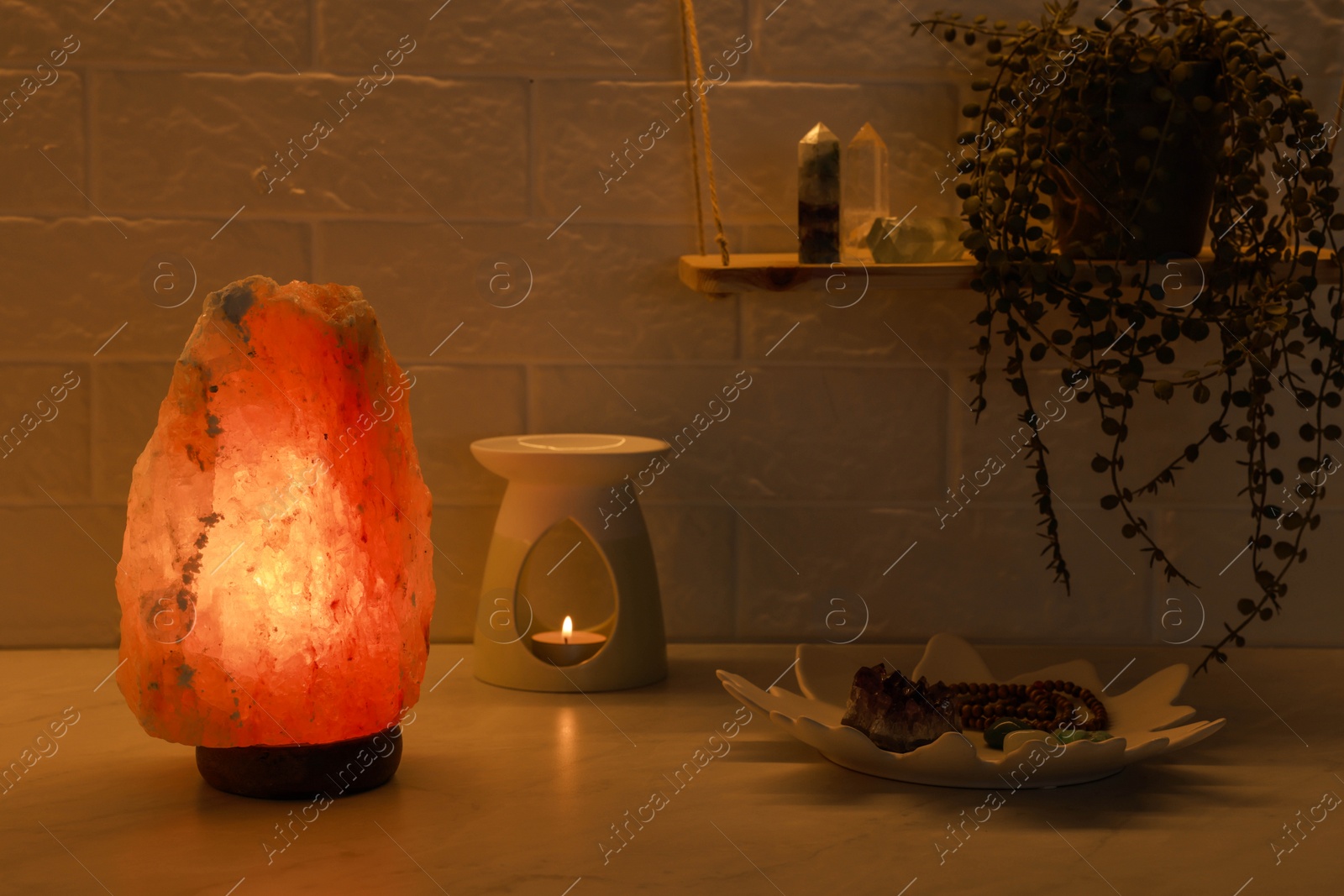 Photo of Himalayan salt lamp, crystals and oil diffuser on white table near brick wall