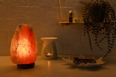 Photo of Himalayan salt lamp, crystals and oil diffuser on white table near brick wall