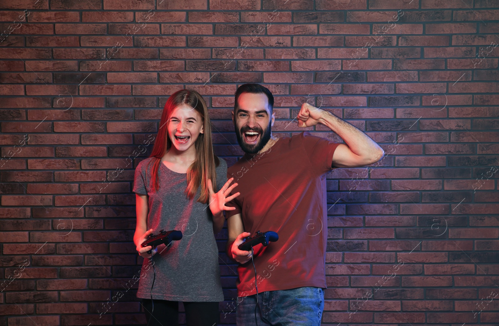 Photo of Young man and teenage girl playing video games with controllers near brick wall