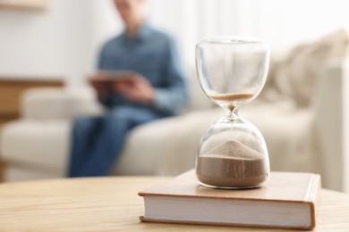 Hourglass with flowing sand on desk. Man reading book in room, selective focus