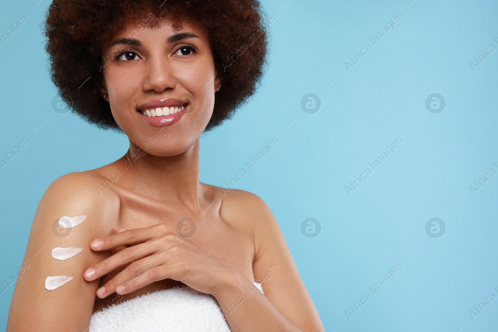 Photo of Beautiful young woman applying body cream onto arm on light blue background, space for text