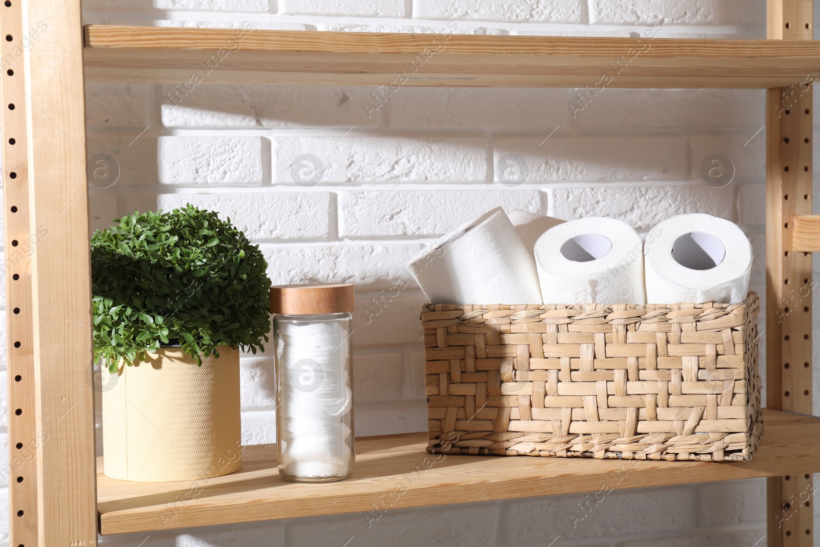 Photo of Toilet paper rolls in wicker basket, floral decor and cotton pads on wooden shelf near white brick wall