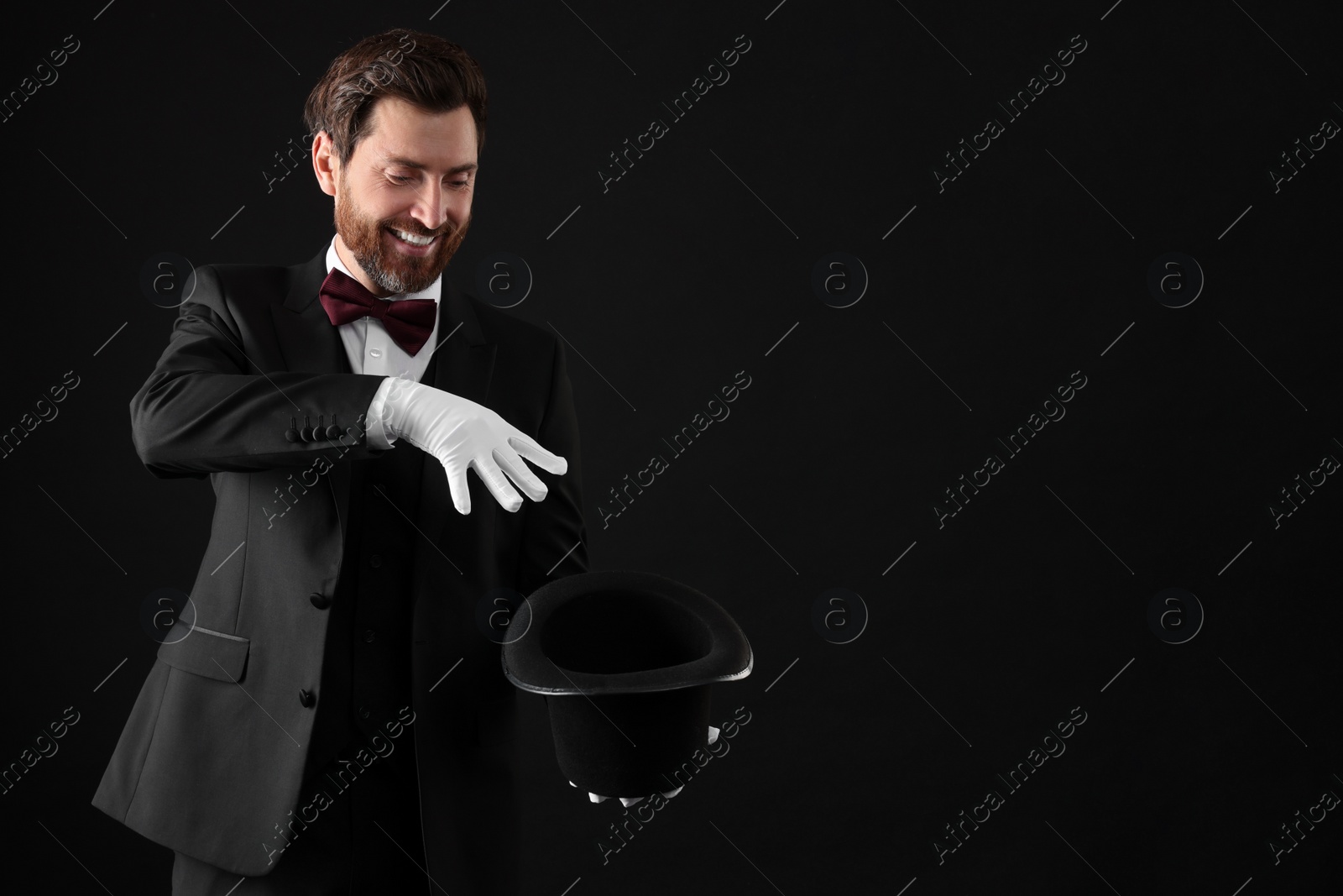 Photo of Happy magician showing magic trick with top hat on black background, space for text