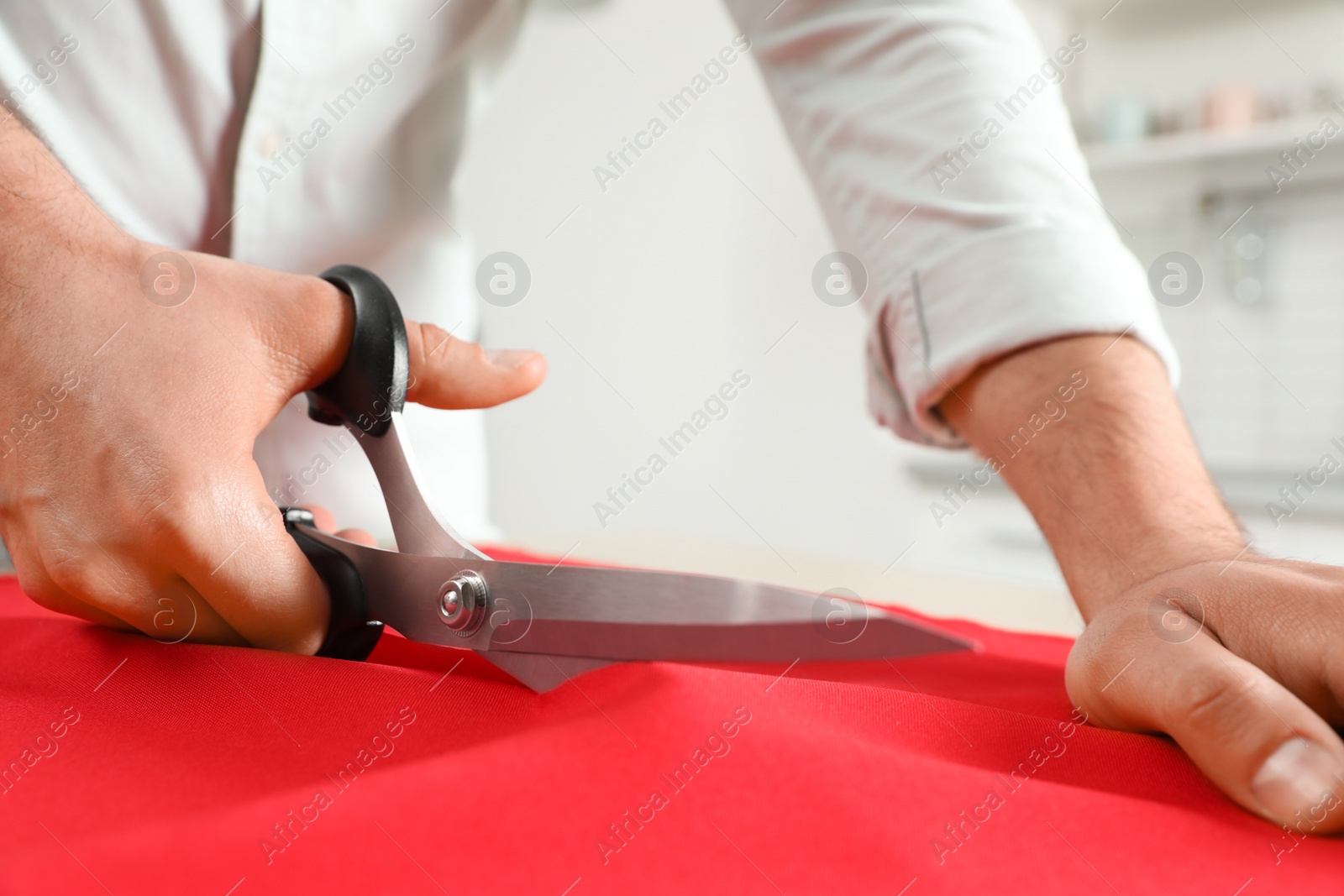 Photo of Professional tailor cutting red fabric with scissors in workshop, closeup