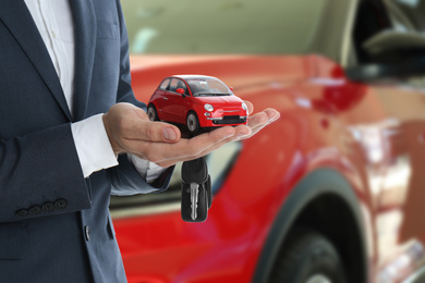 Car buying. Man holding key and model against blurred automobile, closeup
