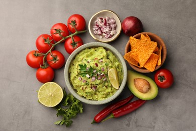 Photo of Bowl of delicious guacamole, nachos chips and ingredients on grey table, flat lay