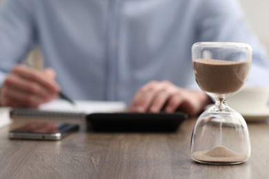 Photo of Hourglass with flowing sand on desk. Man taking notes while using calculator indoors, selective focus